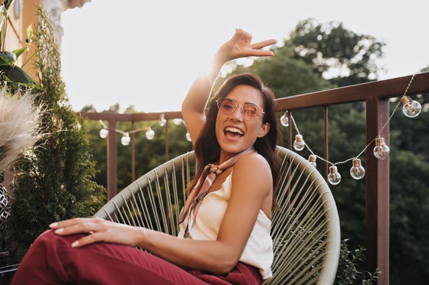 Brunette girl in pink glasses poses on balcony and shows peace sign. cheerful woman in silk scarf around her neck laughs and sits on terrace. Free Photo