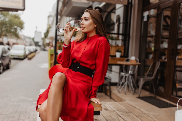 Gorgeous lady in expensive designer dress drinks delicious sparkling wine from crystal glass. full-length shot of blogger sitting in cafe Free Photo