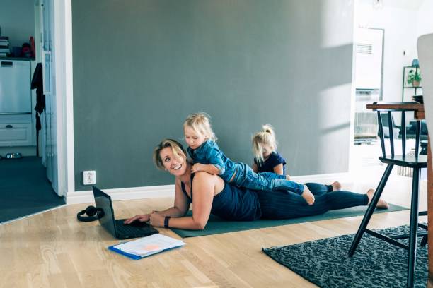 smiling mother with laptop looking at daughter lying on her back while girl sitting in living room at home - mum with kid stock pictures, royalty-free photos & images