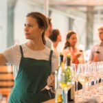 Female wine tasting room worker behind a bar and holding a glass of white wine up to examine it. Guests are laughing in the background.