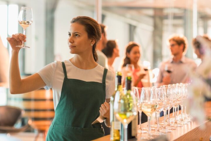 Female wine tasting room worker behind a bar and holding a glass of white wine up to examine it. Guests are laughing in the background.