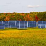 A small field surrounded by autumn-colored trees with a sparse solar farm consisting of nine, 24-paneled solar panels.