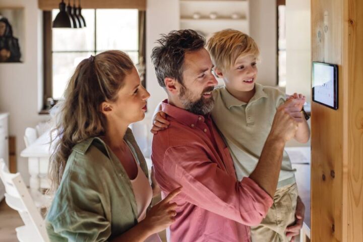 A mother stands next to her husband and son. The father holds the boy and teaches him to use a smart thermostat in the house.